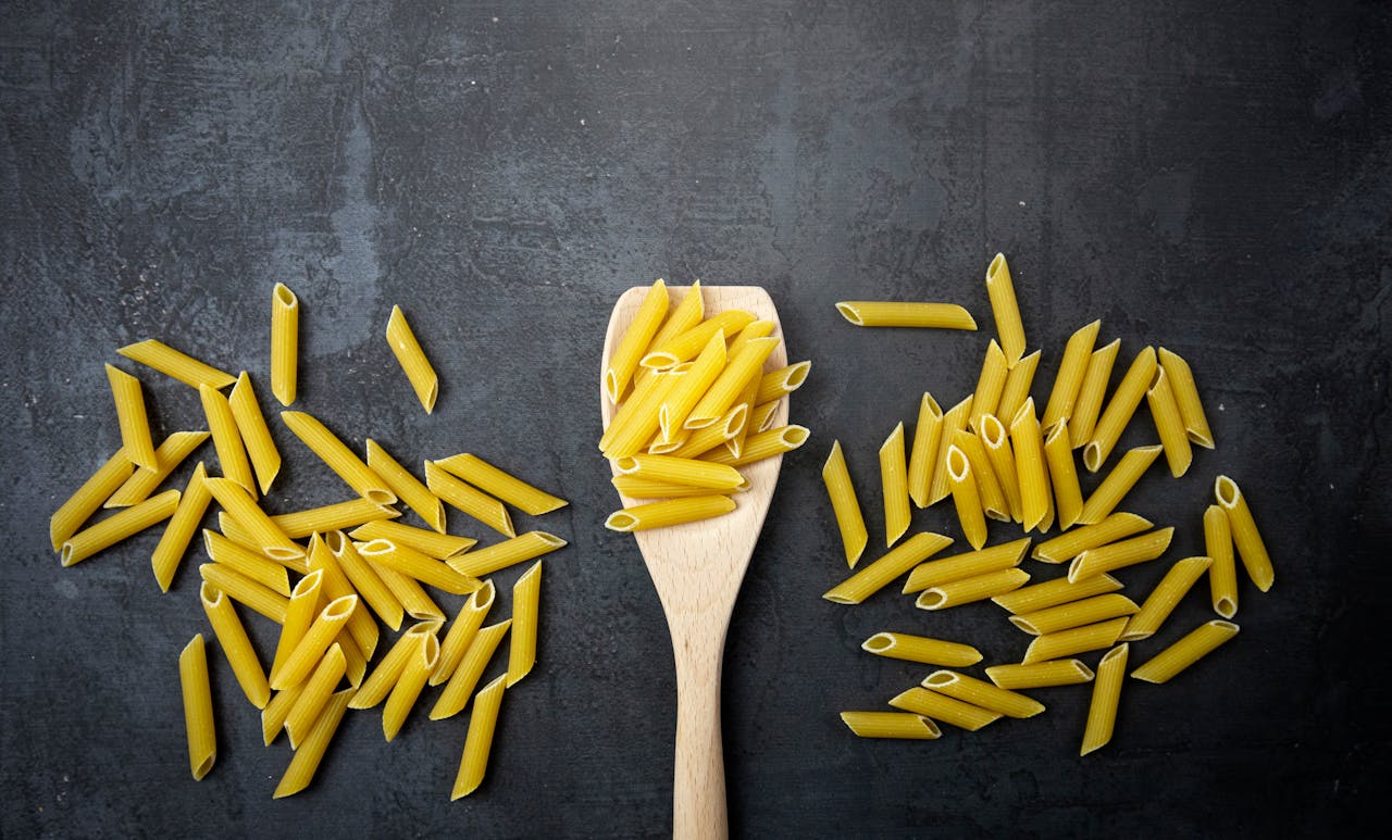 Top view of uncooked penne pasta and wooden spoon on a black surface, perfect for food-related content.