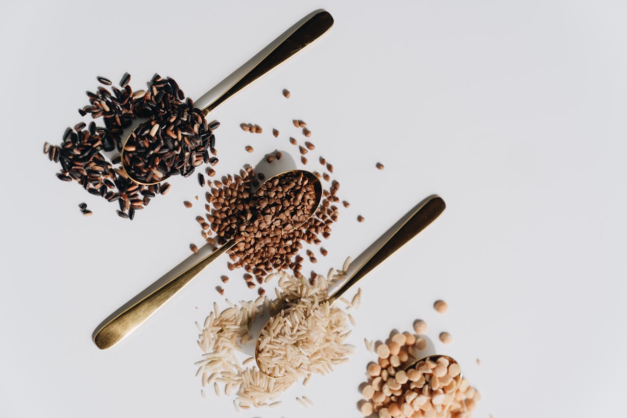 Top view of various dry grains on spoons against a white background, showcasing a variety of textures and colors.