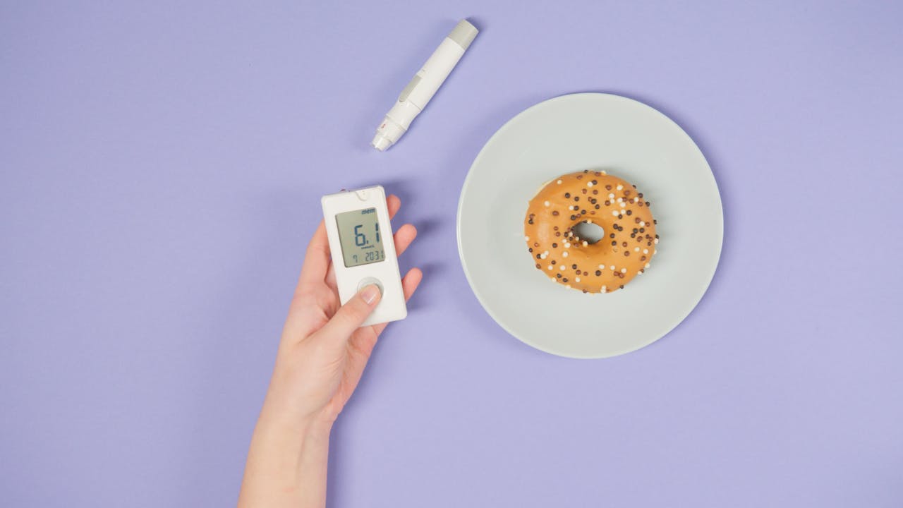 Hand holding a glucometer next to a doughnut on a pastel plate, symbolizing diabetes awareness.